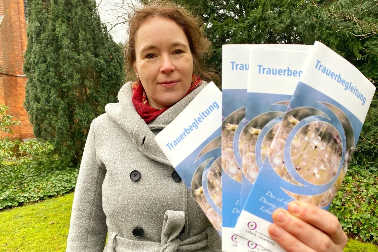 Eine Frau steht auf einem Friedhof, schaut in die Kamera und hält Flyer in der Hand. 